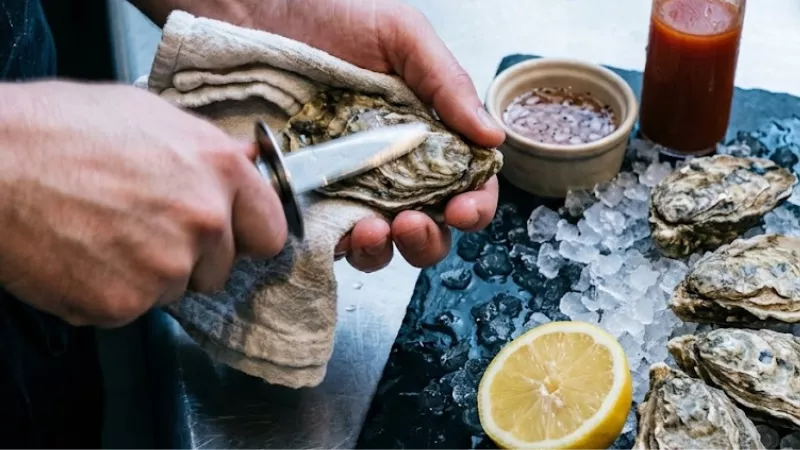 Hands shucking fresh oysters with a knife at the raw bar inside Shuck Me restaurant in Hochatown