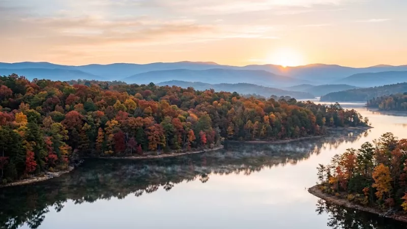 Aerial sunrise view of fall foliage along Broken Bow Lake near Hochatown Oklahoma in late October showing peak red, orange, and golden colors with morning mist over the water and Ouachita Mountain ridges in the background